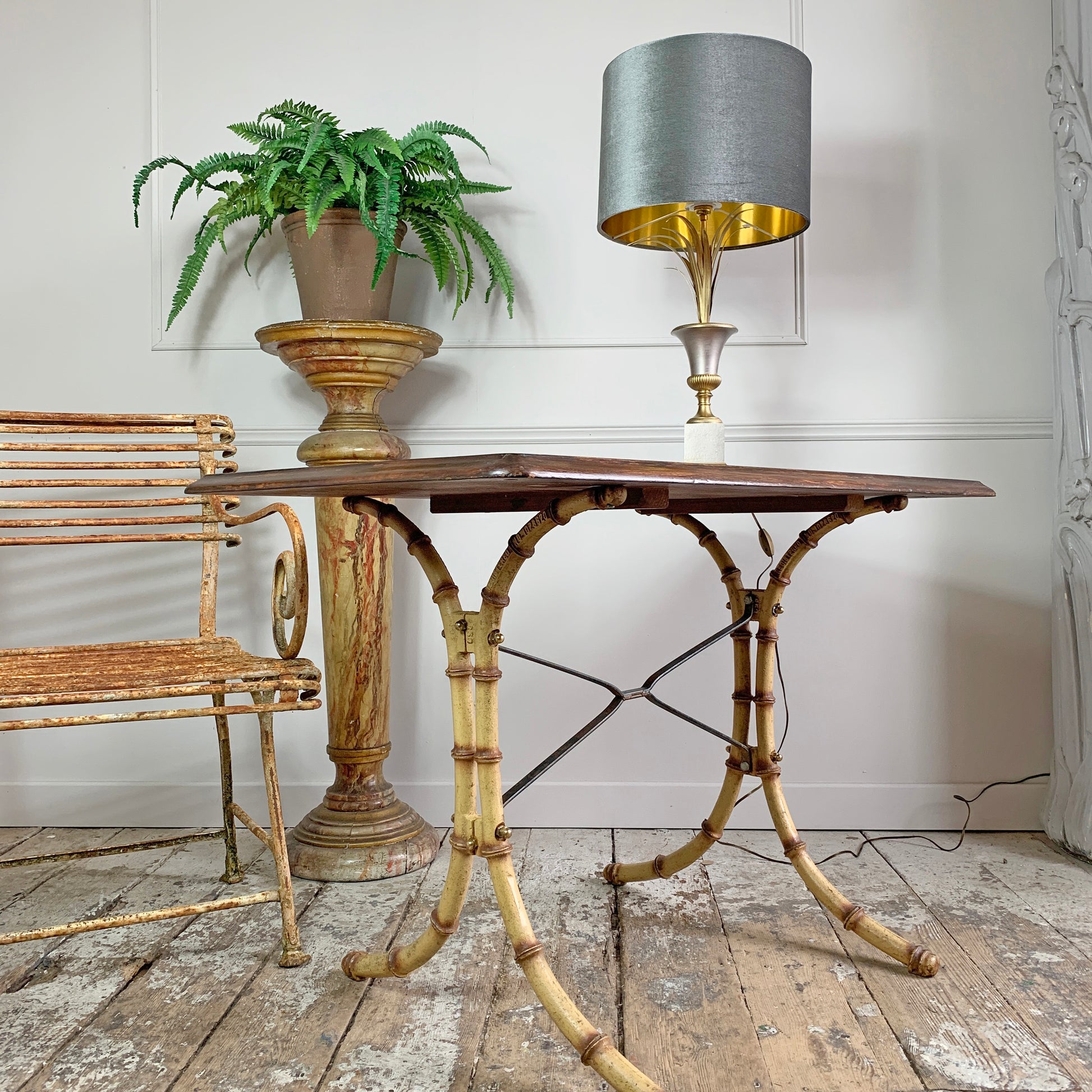 A vintage simulated bamboo table with a dark wood top, styled in a rustic room next to an arras armchair, a potted fern on a pedestal, and a table lamp with a gray and gold shade.