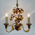 A decorative three-light chandelier with a gold leaf and lemon motif, featuring dark brown wooden arms and candle-style bulbs with illuminated glass shades, set against a plain light gray wall.