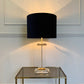 A table lamp with a blue fabric drum shade, a clear glass and brass metal base, and a warm yellow light glowing from within, set on a glass-topped side table against a white paneled wall.