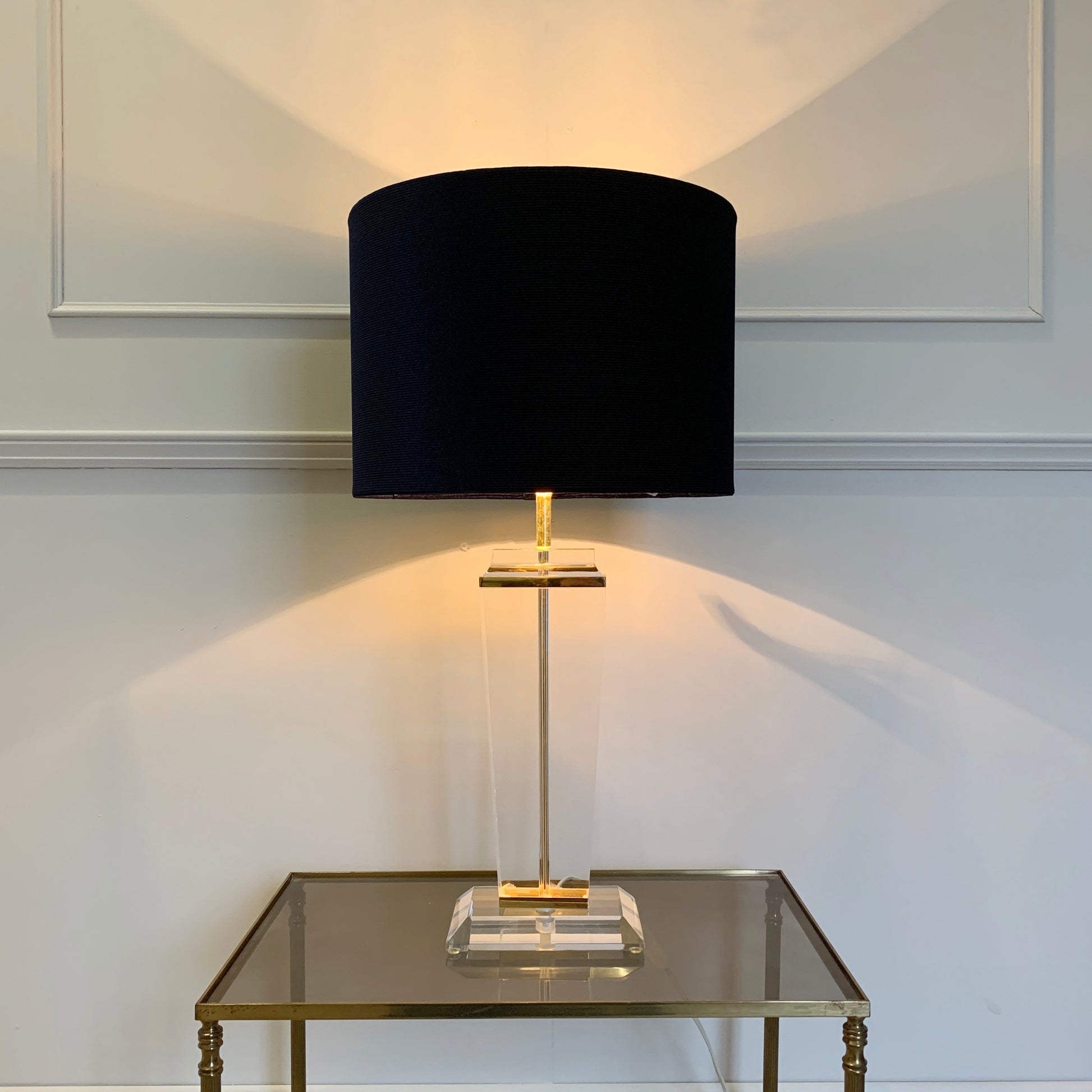 A table lamp with a blue fabric drum shade, a clear glass and brass metal base, and a warm yellow light glowing from within, set on a glass-topped side table against a white paneled wall.