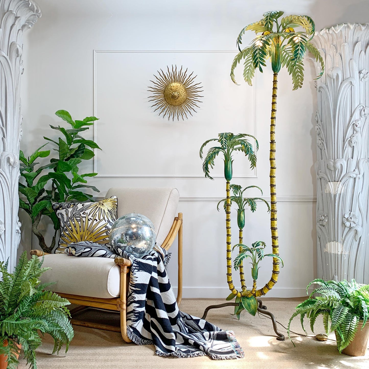 A styled interior corner featuring a tall decorative palm tree with yellow-green trunks and green leaves, alongside a beige armchair with a black-and-white striped throw, potted ferns, and white carved wall panels.