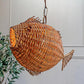 A mid century fish-shaped woven pendant light with a warm orange glow from within, hanging against a white panelled wall with a green plant visible on the left.