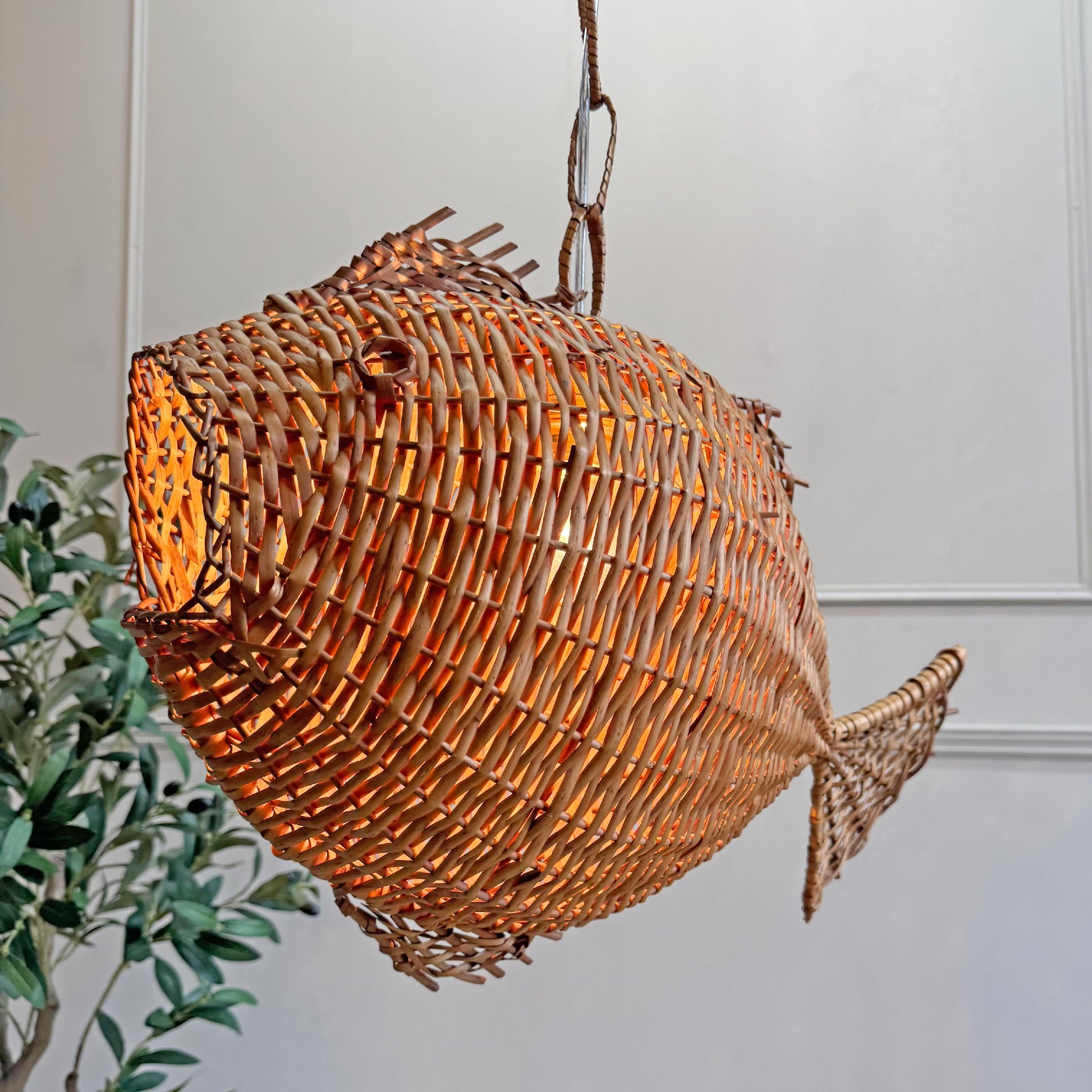 A mid century fish-shaped woven pendant light with a warm orange glow from within, hanging against a white panelled wall with a green plant visible on the left.