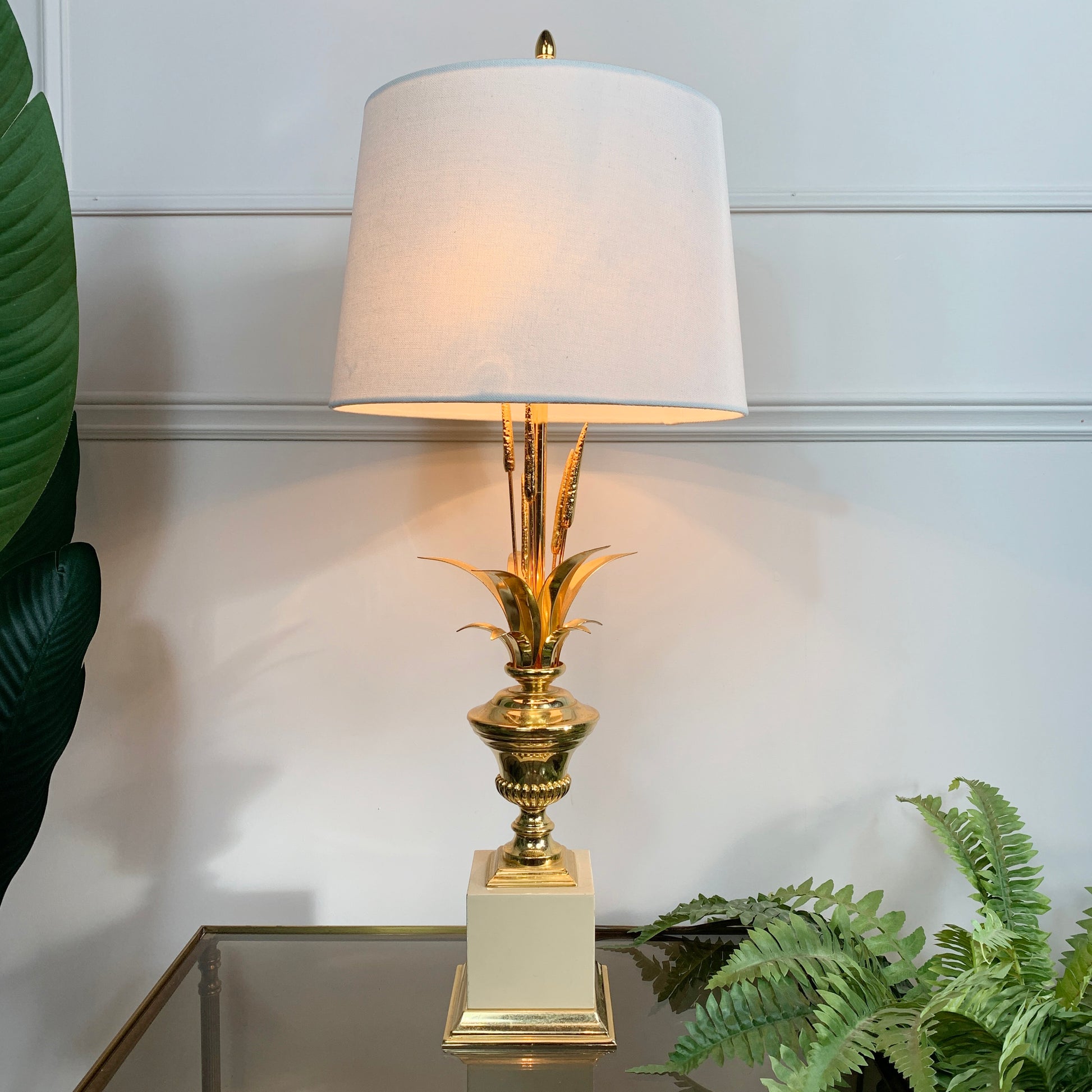 A table lamp with a white drum shade, a gold pineapple-shaped stem base, and a cream-colored square pedestal, shown lit on a glass tabletop with green plants in the background.