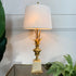 A table lamp with a white drum shade, a gold pineapple-shaped stem base, and a cream-colored square pedestal, shown lit on a glass tabletop with green plants in the background.