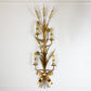 A vintage wall sconce with a gold-toned metal frame shaped like reeds or wheat stalks, featuring multiple candle-style bulbs and a bow detail at the base, mounted on a plain white wall.