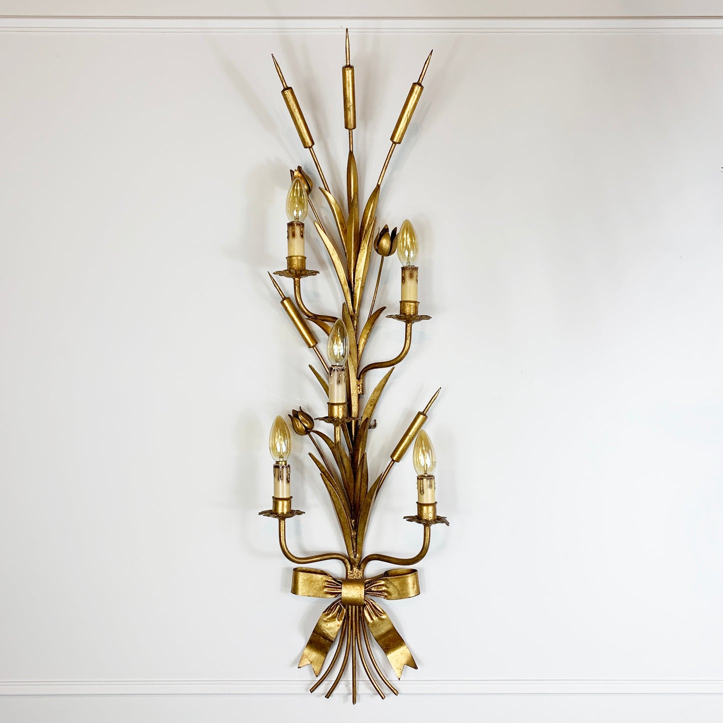 A vintage wall sconce with a gold-toned metal frame shaped like reeds or wheat stalks, featuring multiple candle-style bulbs and a bow detail at the base, mounted on a plain white wall.