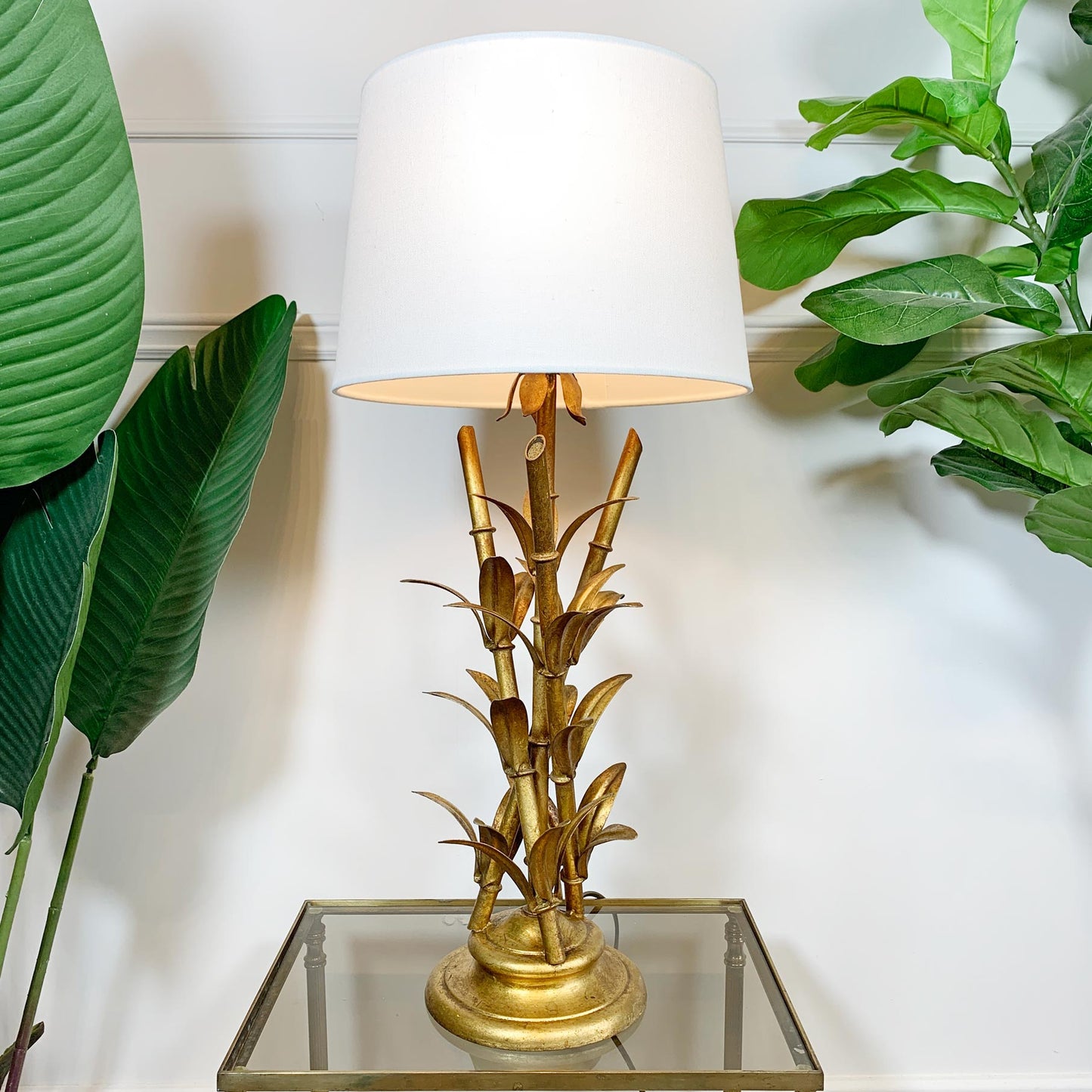 A table lamp with a white drum shade and a gold-toned base sculpted in the shape of bamboo stalks and leaves, displayed on a glass-top side table against a white paneled wall with green plants on both sides.