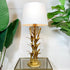 A table lamp with a white drum shade and a gold-toned base sculpted in the shape of bamboo stalks and leaves, displayed on a glass-top side table against a white paneled wall with green plants on both sides.