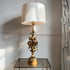 A table lamp with a large white drum shade and an ornate gold-toned sculptural base, shown lit on a glass-topped side table against a light gray wall.