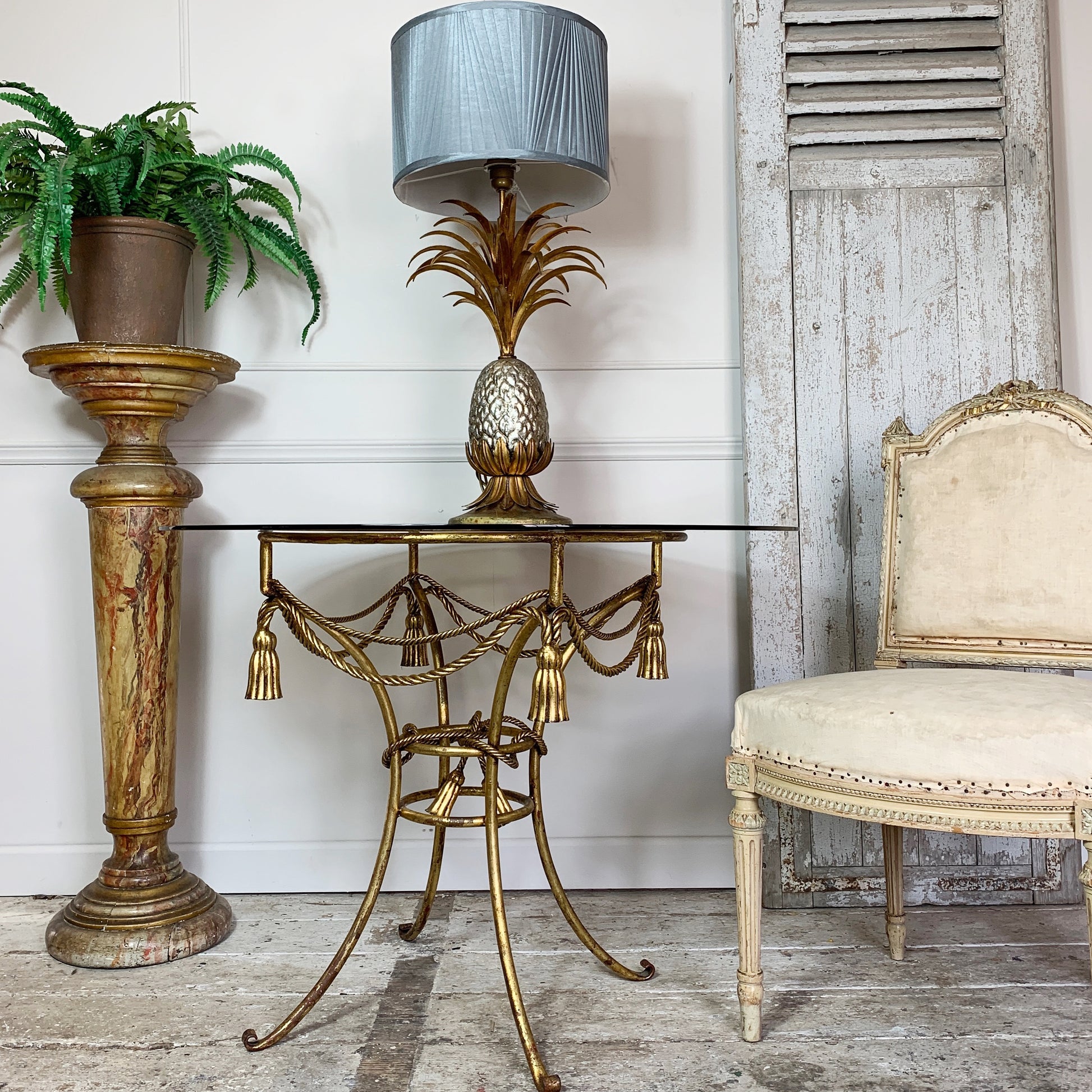 A gold metal table with a glass top and tassel details, styled with a pineapple-shaped table lamp that has a blue-gray pleated shade. The table is set in a rustic room next to a distressed white chair and a potted fern on a marble-patterned pedestal.