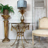 A gold metal table with a glass top and tassel details, styled with a pineapple-shaped table lamp that has a blue-gray pleated shade. The table is set in a rustic room next to a distressed white chair and a potted fern on a marble-patterned pedestal.