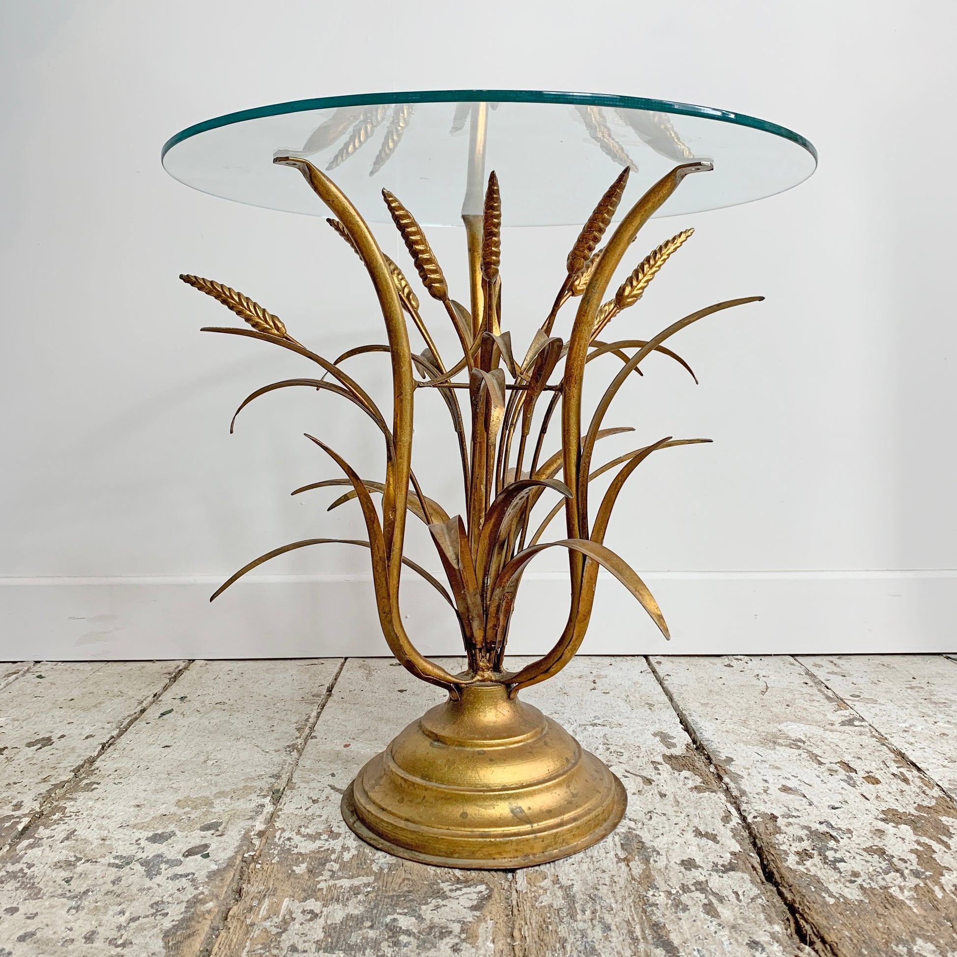 A vintage gold side table featuring a sculptural metallic base resembling wheat stalks in a gold finish, supporting a round clear glass tabletop, set on a rustic white-washed wooden floor against a white wall.