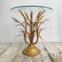 A vintage gold side table featuring a sculptural metallic base resembling wheat stalks in a gold finish, supporting a round clear glass tabletop, set on a rustic white-washed wooden floor against a white wall.