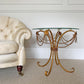 An ornate vintage gold metal accent table with a curved scrollwork base, draped bead chains, and tassels, topped with a clear glass surface. It is pictured in a styled room setting next to a cream tufted armchair on a beige carpet.