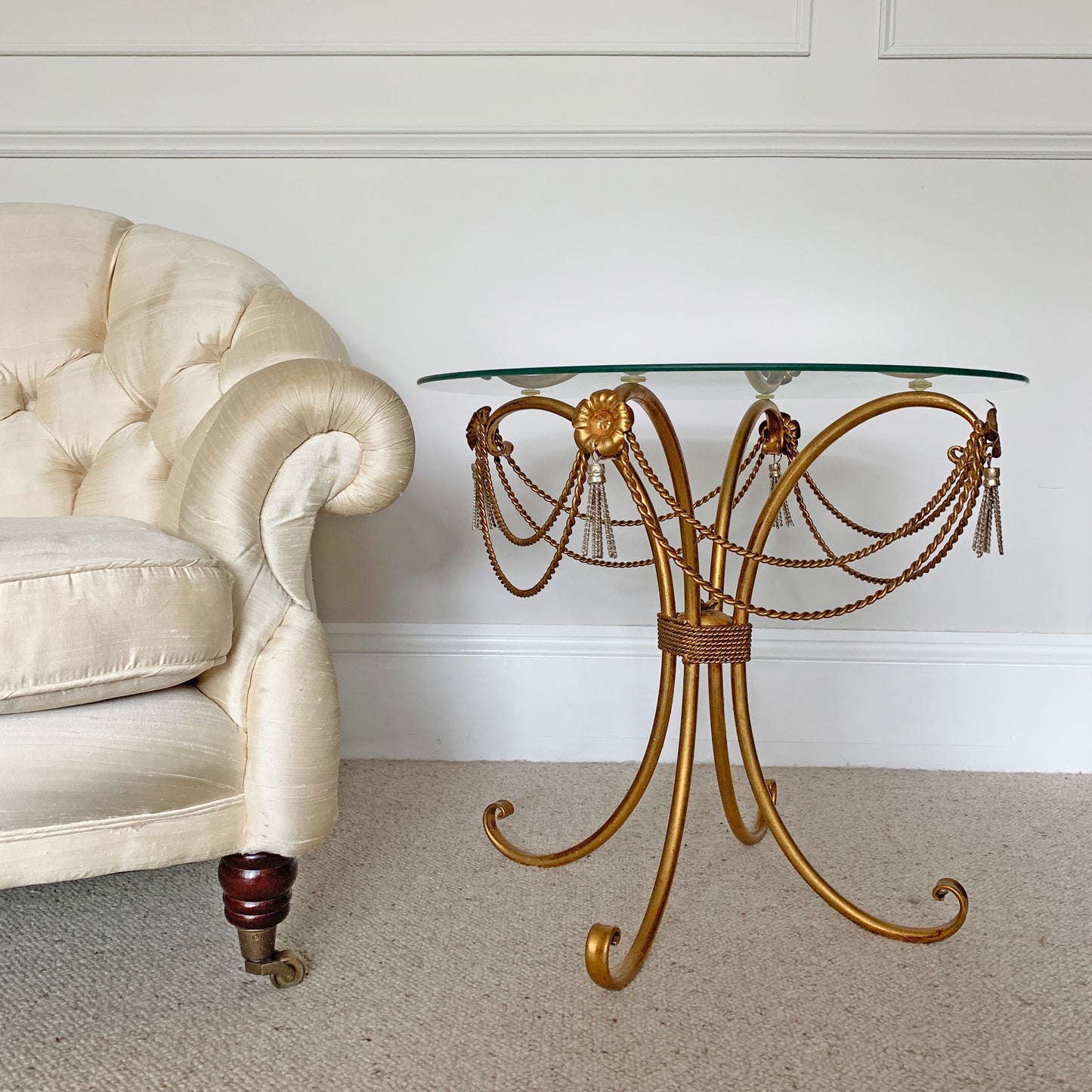 An ornate vintage gold metal accent table with a curved scrollwork base, draped bead chains, and tassels, topped with a clear glass surface. It is pictured in a styled room setting next to a cream tufted armchair on a beige carpet.