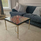 A square fibre optic coffee table with a gold metal frame and a glass top featuring a red and orange circular pattern, set in a living room next to a dark blue sofa.