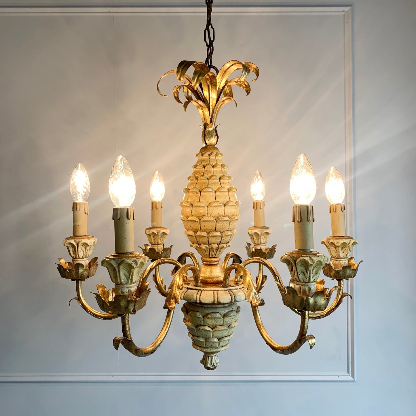 An ornate pineapple-shaped chandelier with gold finish and gilt gold metal accents, shown lit against a white paneled wall.