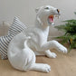 A glossy white ceramic tiger figurine of a roaring big cat, shown sitting on a beige textured surface with a striped pillow and green plant in the background.
