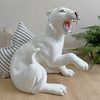 A glossy white ceramic tiger figurine of a roaring big cat, shown sitting on a beige textured surface with a striped pillow and green plant in the background.