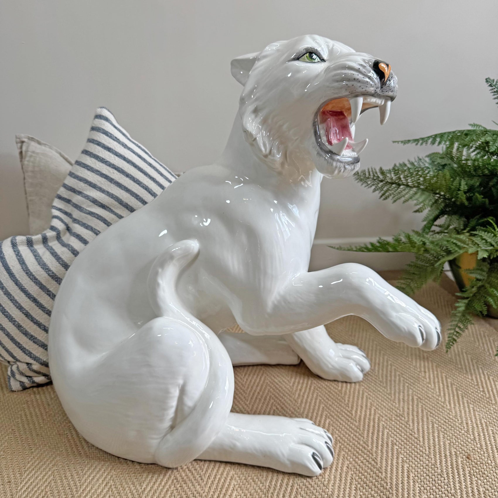 A glossy white ceramic tiger figurine of a roaring big cat, shown sitting on a beige textured surface with a striped pillow and green plant in the background.