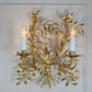 A decorative Italian gold wall sconce with a gold leafy branch design and two lit candle-style bulbs, mounted against a white wall.