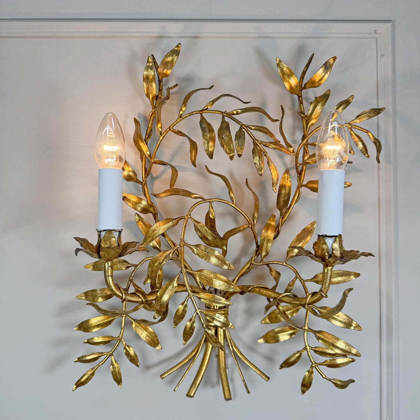 A decorative Italian gold wall sconce with a gold leafy branch design and two lit candle-style bulbs, mounted against a white wall.