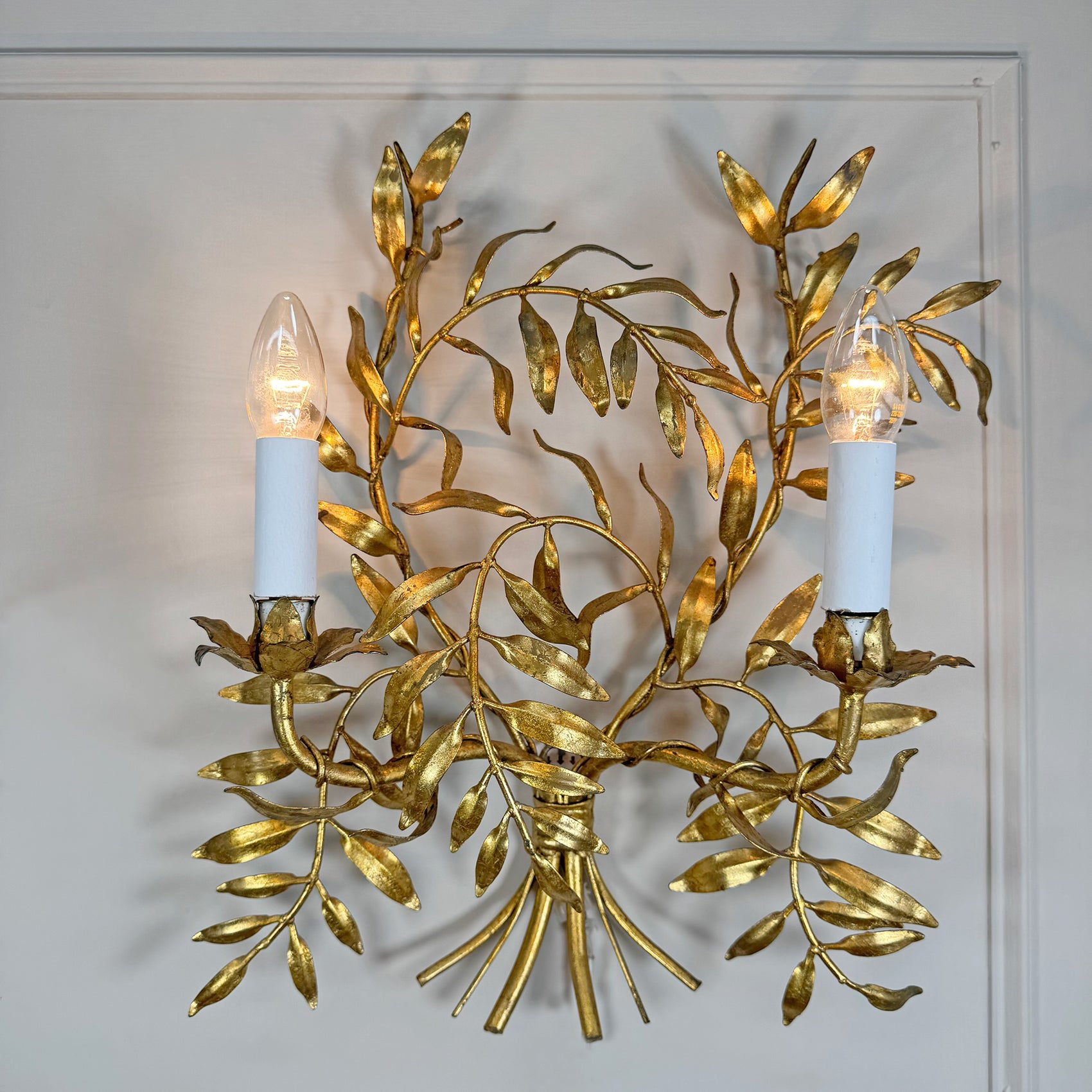 A decorative Italian gold wall sconce with a gold leafy branch design and two lit candle-style bulbs, mounted against a white wall.