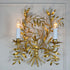A decorative Italian gold wall sconce with a gold leafy branch design and two lit candle-style bulbs, mounted against a white wall.