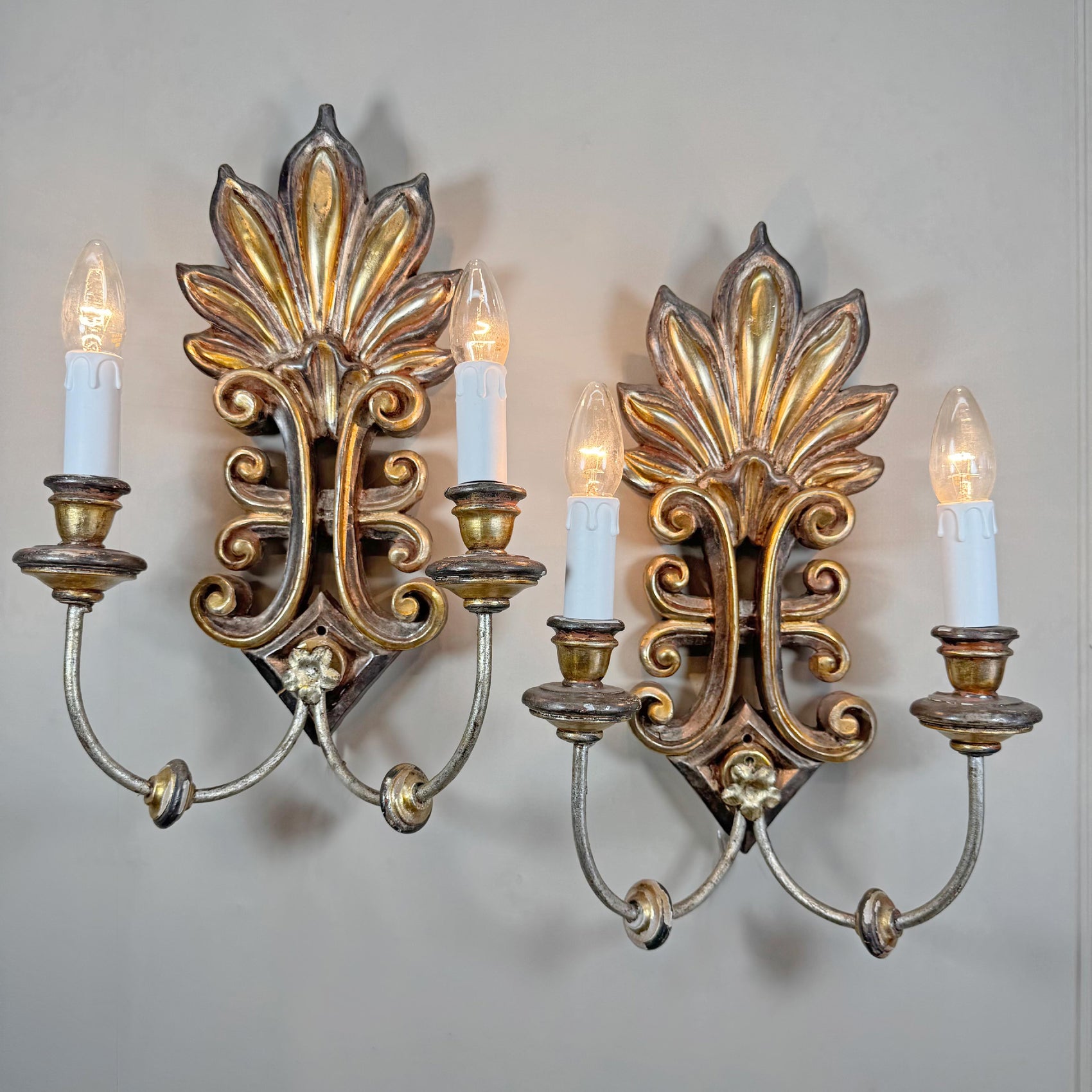 A pair of ornate Italian wooden wall sconces with gilded and antiqued metal scrollwork, each holding two candle-style bulbs, mounted on a plain light gray wall.