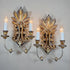 A pair of ornate Italian wooden wall sconces with gilded and antiqued metal scrollwork, each holding two candle-style bulbs, mounted on a plain light gray wall.