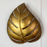 A decorative maison jansen vintage wall-mounted monstera  leaf-shaped sconce in a metallic gold finish with cutout leaf veins, shown against a plain white wall.