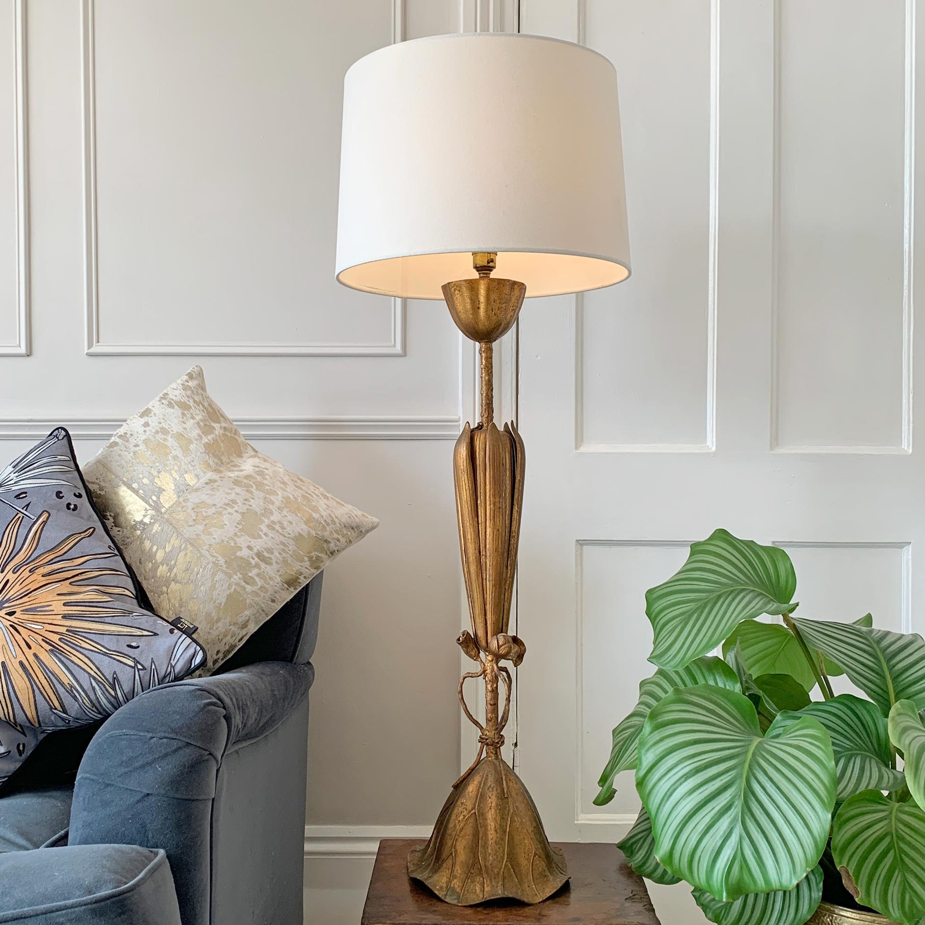 A tall table lamp with a white drum shade and an ornate gold-toned sculptural base, styled in a living room next to a blue armchair and a green plant.
