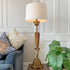 A tall table lamp with a white drum shade and an ornate gold-toned sculptural base, styled in a living room next to a blue armchair and a green plant.