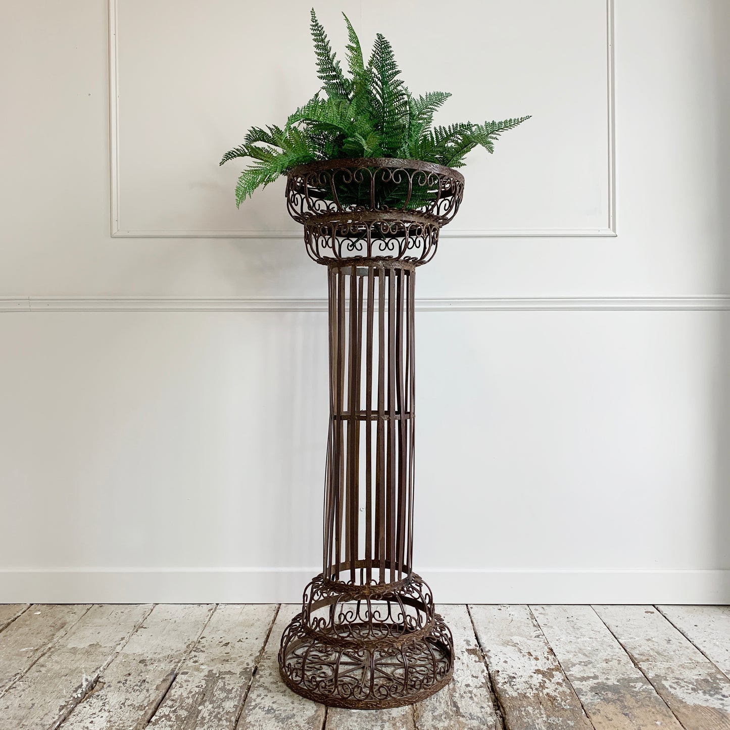 A dark brown ornate metal plant stand with a tiered column design, holding a green fern, set against a white paneled wall and rustic wooden floor.