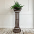A dark brown ornate metal plant stand with a tiered column design, holding a green fern, set against a white paneled wall and rustic wooden floor.