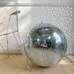A large decorative vintage mirror disco ball made of mirrored tiles sits on a woven beige rug next to a distressed white metal chair, with sunlight casting star-like reflections on a white paneled wall.