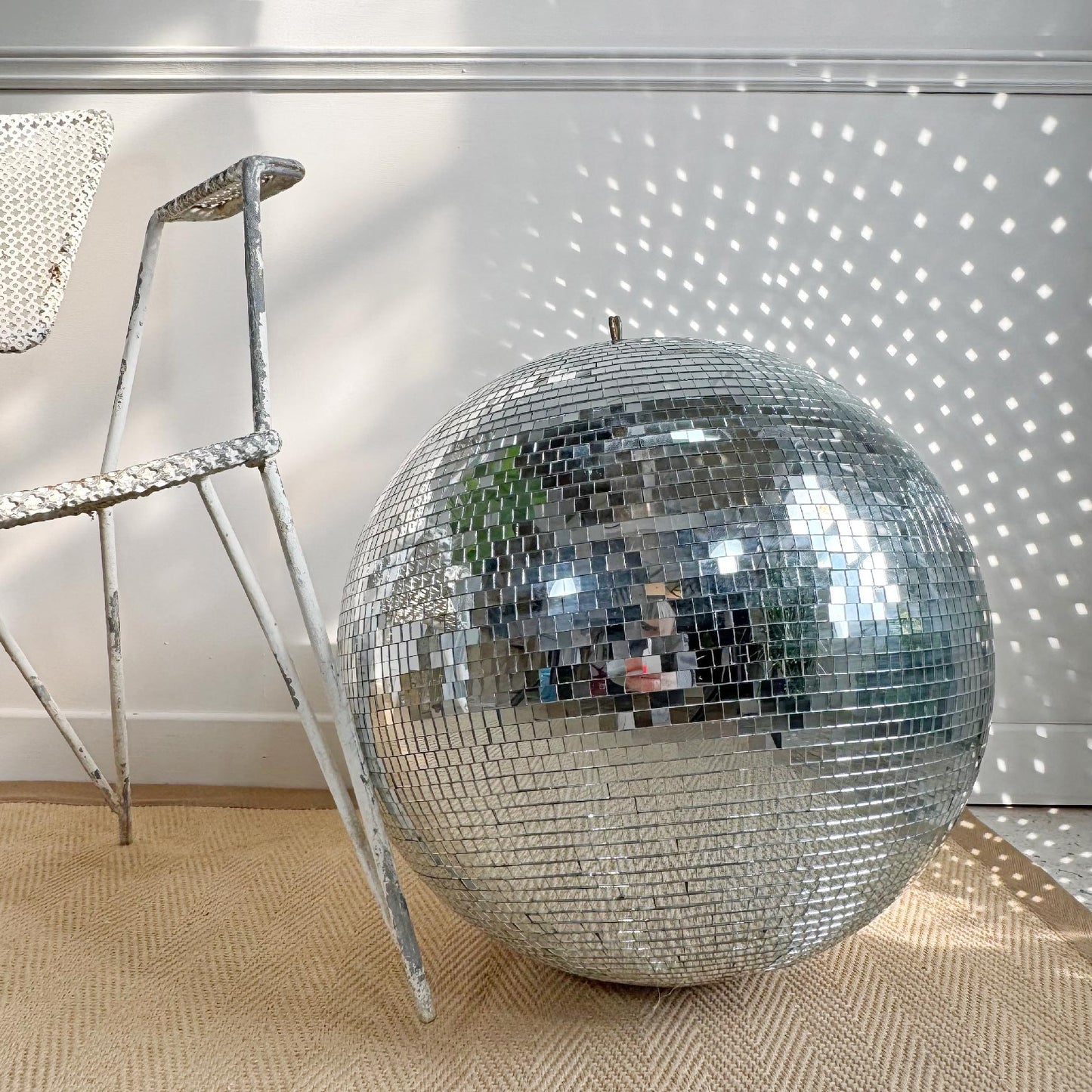 A large decorative vintage mirror disco ball made of mirrored tiles sits on a woven beige rug next to a distressed white metal chair, with sunlight casting star-like reflections on a white paneled wall.