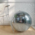 A large decorative vintage mirror disco ball made of mirrored tiles sits on a woven beige rug next to a distressed white metal chair, with sunlight casting star-like reflections on a white paneled wall.