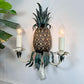 A decorative wall sconce shaped like a pineapple, with a textured brown pineapple body, dark green leafy top and lower details, and two white candle-style holders with lit bulb lights, mounted against a white wall with green leaves entering from the left.