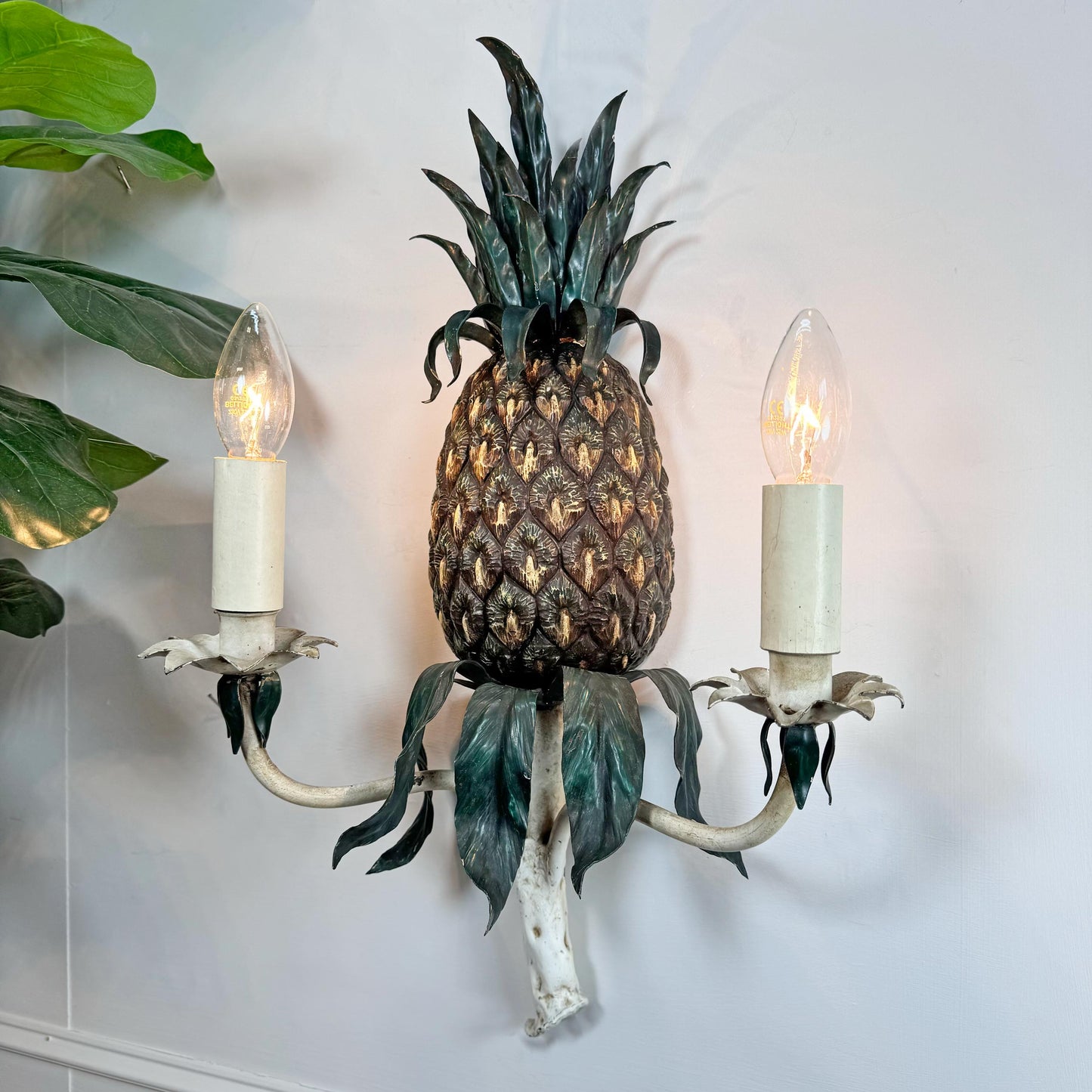 A decorative wall sconce shaped like a pineapple, with a textured brown pineapple body, dark green leafy top and lower details, and two white candle-style holders with lit bulb lights, mounted against a white wall with green leaves entering from the left.