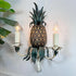 A decorative wall sconce shaped like a pineapple, with a textured brown pineapple body, dark green leafy top and lower details, and two white candle-style holders with lit bulb lights, mounted against a white wall with green leaves entering from the left.