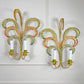 A pair of decorative feather leaf style wall sconces with curved, woven palm-leaf designs in yellow, green, orange, and blue, each holding two white candle-style bulbs, mounted on a white panelled wall.