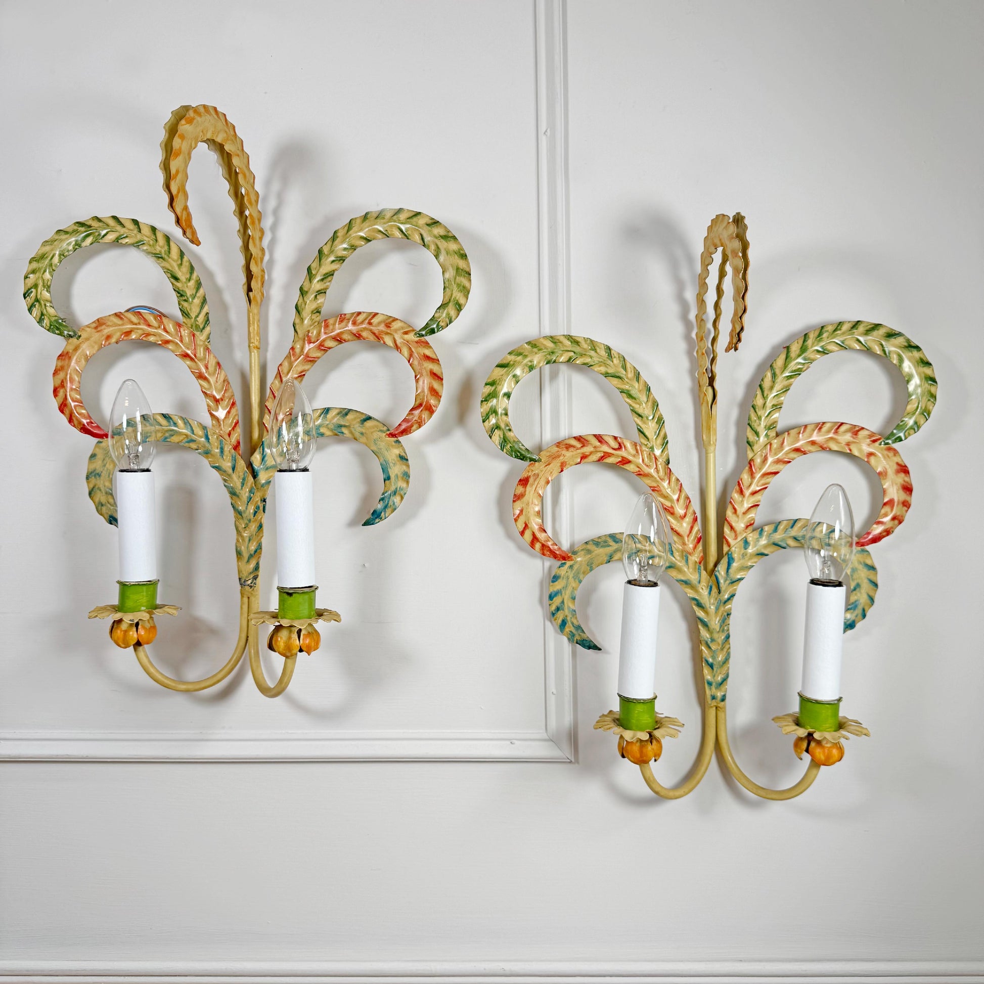 A pair of decorative feather leaf style wall sconces with curved, woven palm-leaf designs in yellow, green, orange, and blue, each holding two white candle-style bulbs, mounted on a white panelled wall.