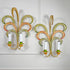 A pair of decorative feather leaf style wall sconces with curved, woven palm-leaf designs in yellow, green, orange, and blue, each holding two white candle-style bulbs, mounted on a white panelled wall.