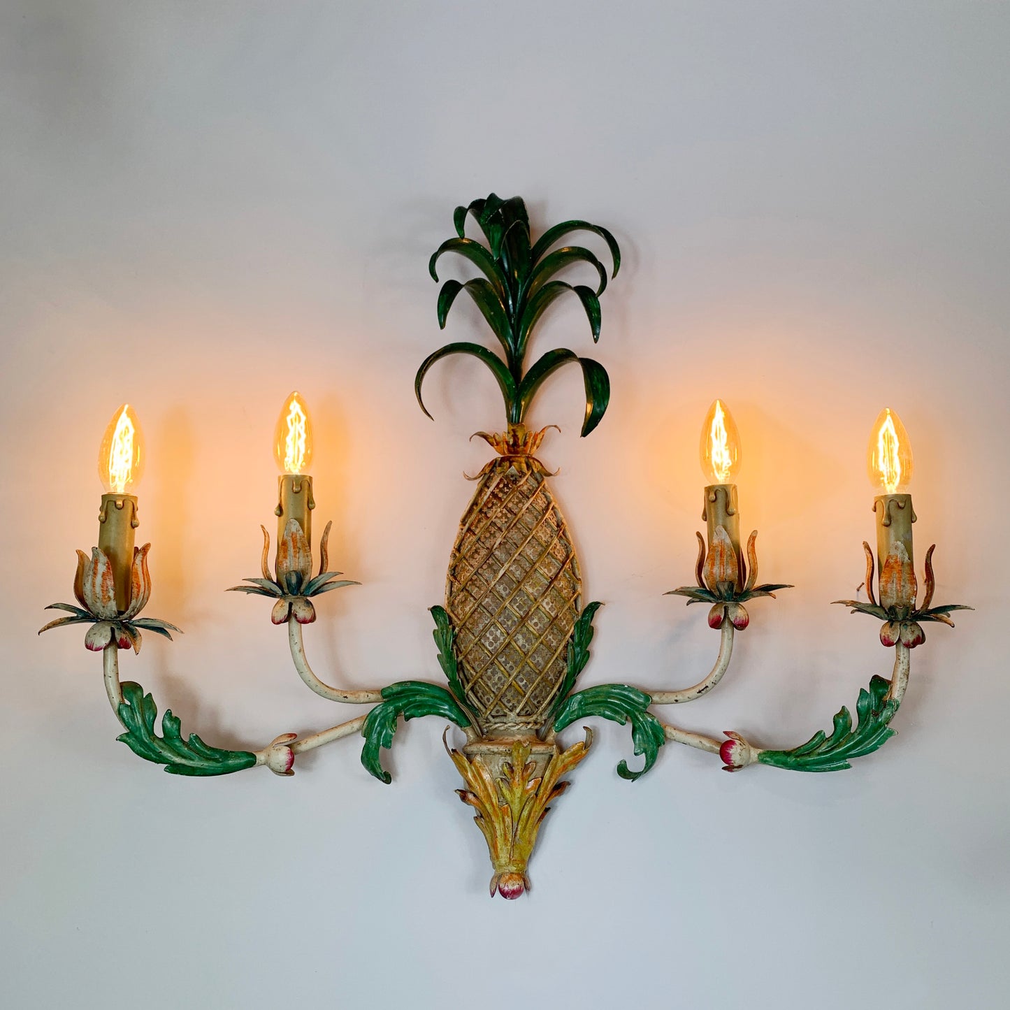A vintage toleware decorative wall sconce shaped like a pineapple, with four lit candle-style bulbs, mounted against a plain light wall.