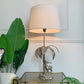 A Valenti of Spain, Stag table lamp with a large beige drum shade, a silver palm tree base, and a deer figurine, shown lit on a glass-top side table with green plants in a bright room.
