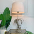 A Valenti of Spain, Stag table lamp with a large beige drum shade, a silver palm tree base, and a deer figurine, shown lit on a glass-top side table with green plants in a bright room.