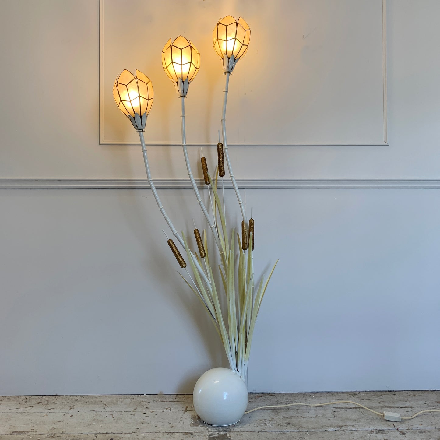 A vintage mid century floor lamp with three glowing geometric flower heads on tall white stems, surrounded by faux cattail leaves and brown seed pods, set in a white round base on a wooden floor against a white wall.
