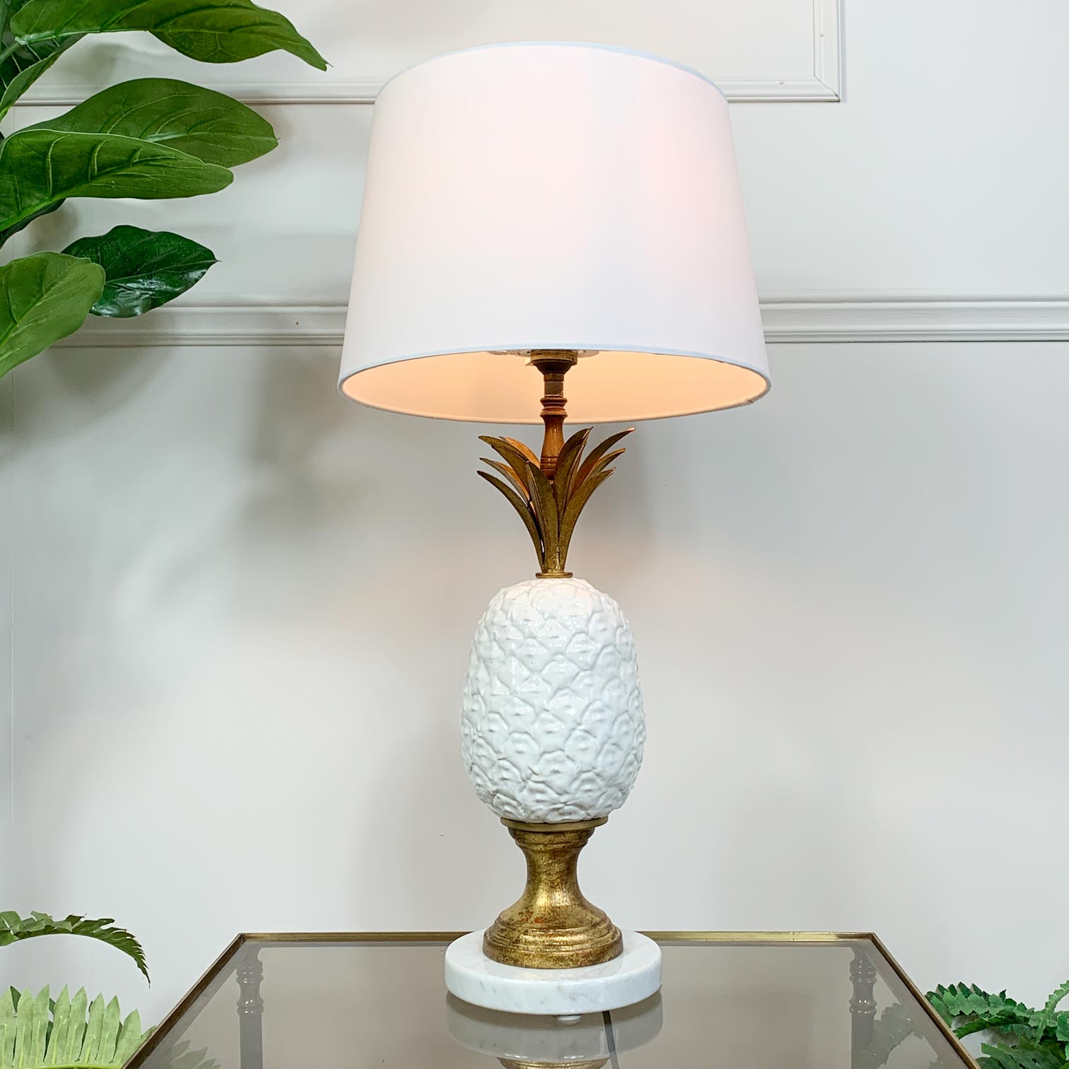 A vintage table lamp shaped like a pineapple, with a ceramic white body, a gold-toned stem and base, and a white drum shade, shown lit on a glass tabletop against a white wall with green plants in the background.